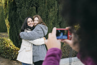 Close-up of young woman photographing female friends embracing in park - ABZF03195