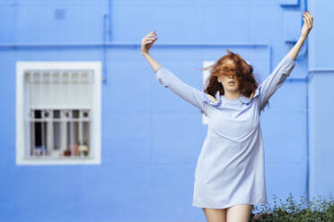 Rothaarige Frau mit erhobenen Armen vor einem blauen Haus - JSMF01553