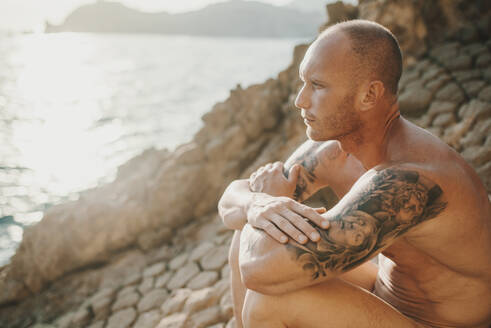 Tattooed nudist sitting on volcanic rocks by the sea - MIMFF00045