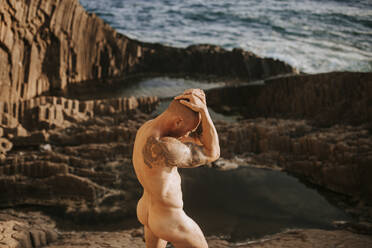 Tätowierter Nudist posiert auf vulkanischen Felsen am Meer - MIMFF00046