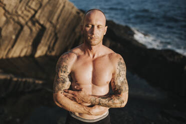 Tattooed nudist standing on volcanic rocks by the sea with arms crossed - MIMFF00047