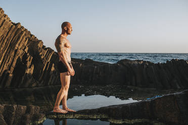 Muscular man in underpants standing on rocks by the sea - MIMFF00049