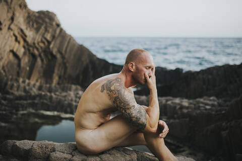 Tätowierter Nudist auf vulkanischen Felsen am Meer sitzend, lizenzfreies Stockfoto