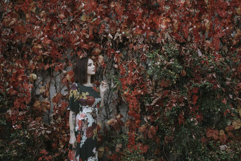 Thoughtful young woman standing against ivy wall during autumn in park stock photo