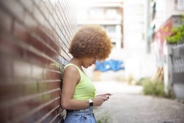 Frau mit Afro-Haar, die ein Smartphone benutzt, während sie an einer Mauer steht - OCMF01349