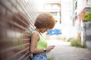 Woman with afro hair using smart phone while standing by brick wall - OCMF01349