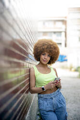 Mid adult woman with afro hair using smart phone while leaning on brick wall in city - OCMF01350