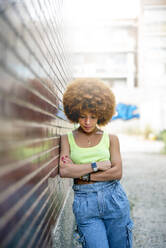 Mittlere erwachsene Frau mit Afro-Haar, die nach unten schaut, während sie an einer Backsteinmauer in der Stadt steht - OCMF01351