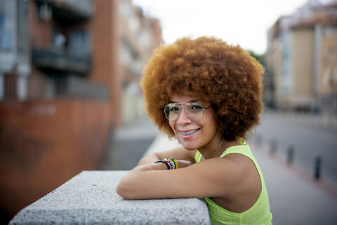 Nahaufnahme einer lächelnden Frau mit Afro-Haar, die an einer Stützmauer in einer Stadt steht, lizenzfreies Stockfoto