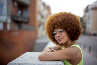 Nahaufnahme einer Frau mit Afro-Haar, die eine Brille trägt und auf einer Stützmauer in der Stadt steht - OCMF01353