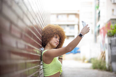 Mittlere erwachsene Frau mit Afro-Haar, die ein Selfie mit ihrem Smartphone macht, während sie an einer Mauer steht - OCMF01355