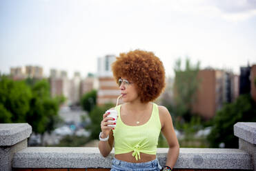 Mittlere erwachsene Frau mit Afro-Haar trinkt Saft, während sie gegen den Himmel in der Stadt steht - OCMF01356