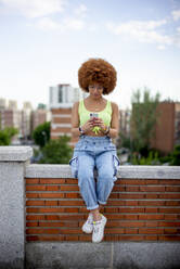 Frau mit Afrofrisur, die ihr Smartphone benutzt, während sie auf einer Stützmauer sitzt - OCMF01358