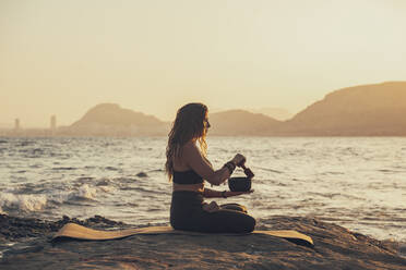 Mature woman meditating with singing bowl practicing yoga at rocky beach in the evening - DLTSF00738