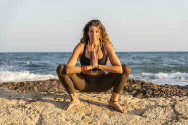 Mature woman practicing yoga at rocky beach in the evening - DLTSF00739