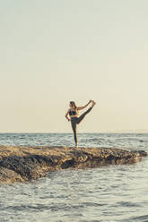 Mature woman practicing yoga at rocky beach in the evening - DLTSF00740