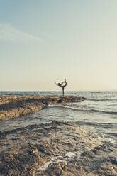 Mature woman practicing yoga at rocky beach in the evening, dancer position - DLTSF00743