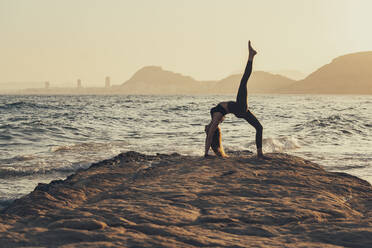 Mature woman practicing yoga at rocky beach in the evening, Urdhva Dhanurasana - DLTSF00746