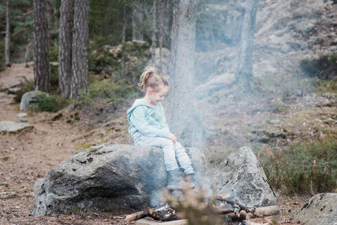 Junges Mädchen sitzt an einem Lagerfeuer und wärmt sich im Wald in Schweden auf - CAVF84984