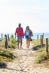 Glückliches Paar mit Sonnenbrille, das sich beim Strandspaziergang an den Händen hält - CAVF84997