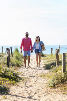 Happy couple with sunglasses holding hands while walking on beach - CAVF84997