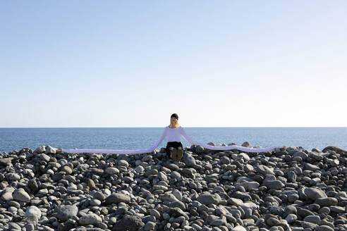 Frau mit künstlichen langen Händen sitzt auf Kieselsteinen am Strand gegen klaren Himmel - PSTF00746