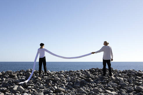 Woman with long artificial arms holding man's hand at beach against clear sky - PSTF00754