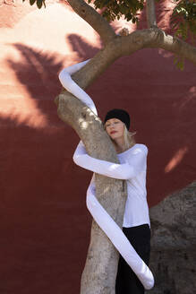 Relaxed woman with artificial long hands embracing tree trunk against wall - PSTF00772