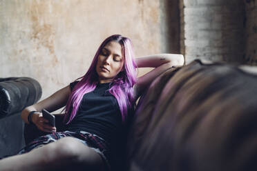 Junge Frau mit rosa Haaren sitzt auf dem Sofa in einem Loft und benutzt ein Smartphone - MEUF00695