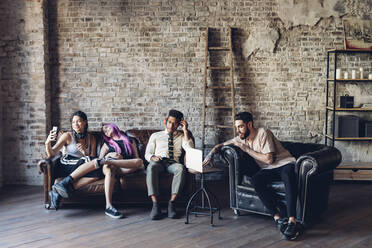 Group of friends sitting on sofa in a loft using technology - MEUF00703