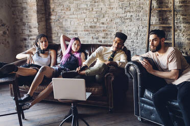 Group of friends sitting on sofa in a loft looking at laptop - MEUF00704