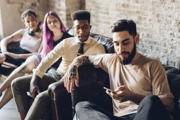 Group of friends sitting on sofa in a loft with smartphone - MEUF00705