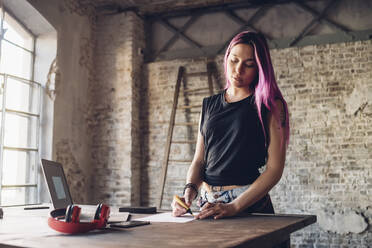 Creative businesswoman with pink hair working at table in loft office - MEUF00737