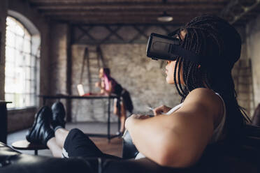 Junge Frau entspannt sich in einem Sessel in einem Loft-Büro und trägt eine VR-Brille - MEUF00740