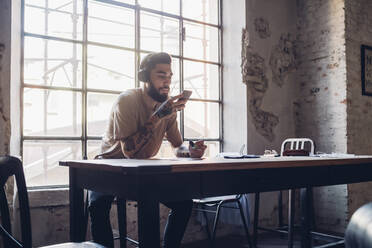 Kreativer junger Geschäftsmann mit Kopfhörern und Smartphone sitzt am Tisch im Loft-Büro - MEUF00741