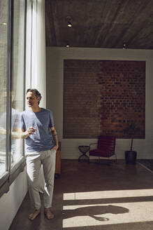 Senior man looking out of window in a loft flat - MCF00929