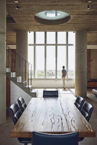 Holztisch in einer Loftwohnung mit Mann am Fenster im Hintergrund - MCF00933