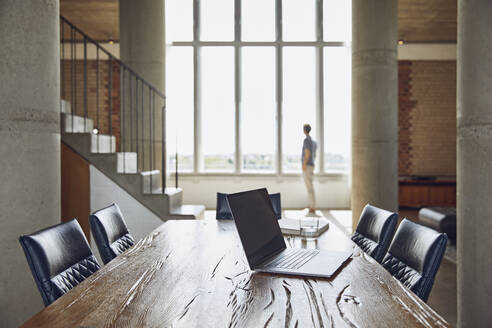 Laptop on wooden table in a loft flat with man at the window in background - MCF00934