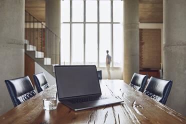 Laptop on wooden table in a loft flat with man at the window in background - MCF00936