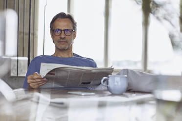 Älterer Mann sitzt am Tisch in einer Dachgeschosswohnung und liest Zeitung - MCF00941