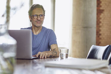 Senior man sitting at the table in a loft flat using laptop - MCF00943
