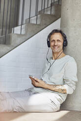 Smiling senior man with headphones listening music in a loft flat - MCF00968