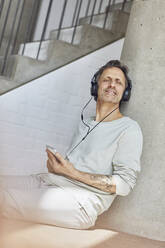 Älterer Mann mit Kopfhörern, der in einer Loftwohnung Musik hört - MCF00969