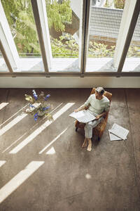 Älterer Mann sitzt auf einem Stuhl in einer Dachgeschosswohnung und liest Zeitung - MCF00993