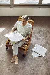 Älterer Mann sitzt auf einem Stuhl in einer Dachgeschosswohnung und liest Zeitung - MCF01000