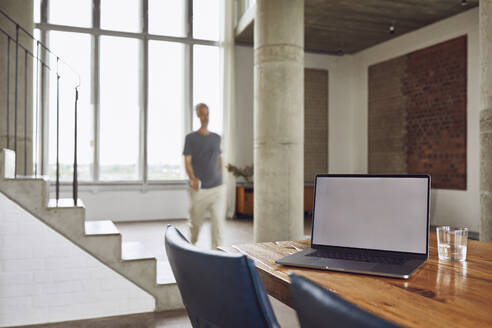 Laptop auf Holztisch in einer Loftwohnung mit Mann im Hintergrund - MCF01026