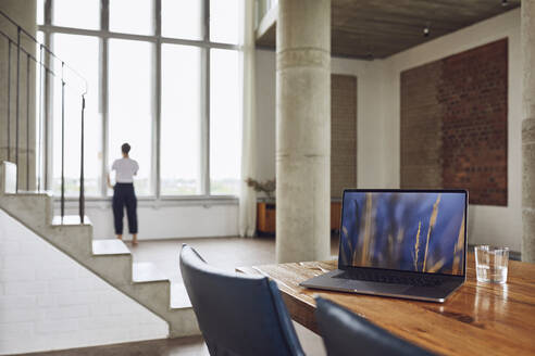 Laptop auf Holztisch in einer Loftwohnung mit Frau am Fenster im Hintergrund - MCF01028