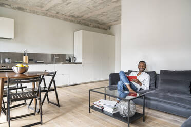 Laughing man with book sitting on couch in modern apartment - AHSF02716