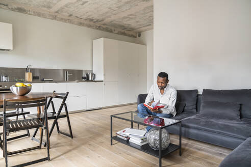 Man sitting on couch in modern apartment turning pages in a book - AHSF02717