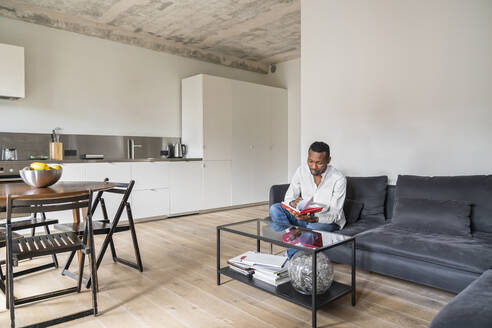 Mann sitzt auf der Couch in einer modernen Wohnung und blättert in einem Buch - AHSF02717